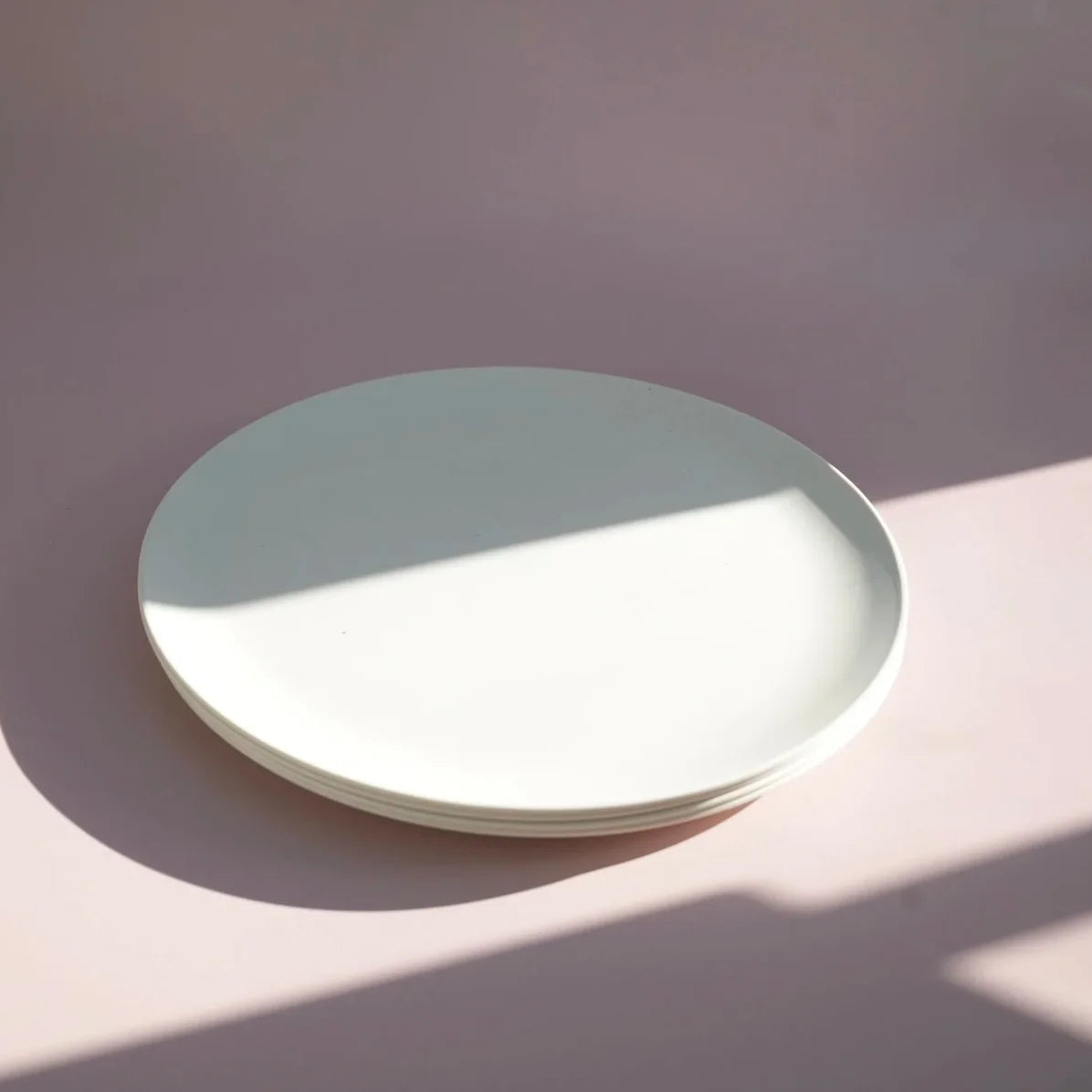 A stack of round white ceramic plates on a dusty pink surface with a diagonal shadow cast across the scene.