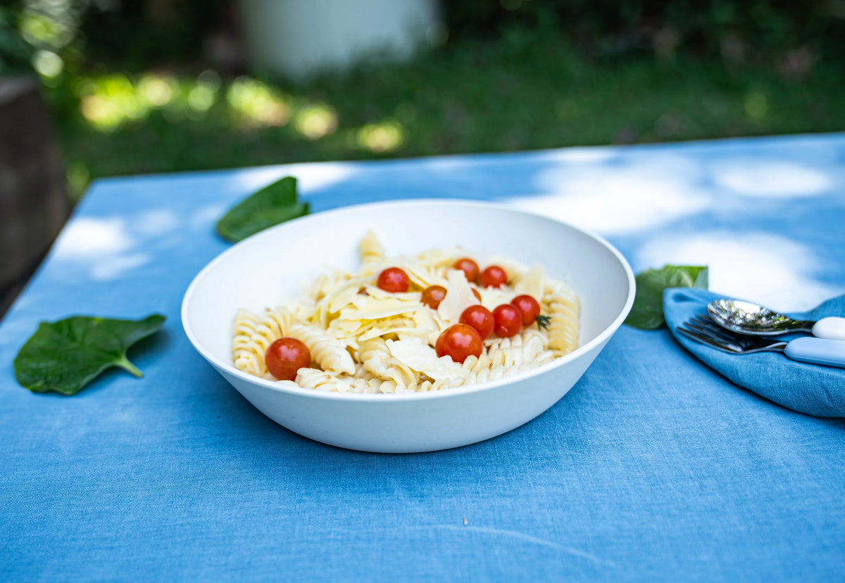 A white bowl of rotini pasta salad with cherry tomatoes, set on a blue tablecloth outdoors with green leaves and a blue napkin with silverware.