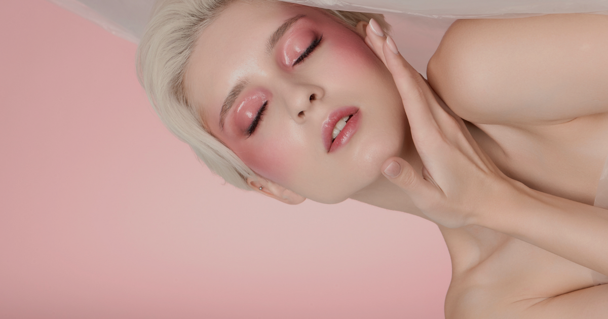 A close-up beauty shot of a woman with short platinum blonde hair, wearing soft pink eyeshadow and blush with glossy pink lips, touching her cheek against a solid pink background.