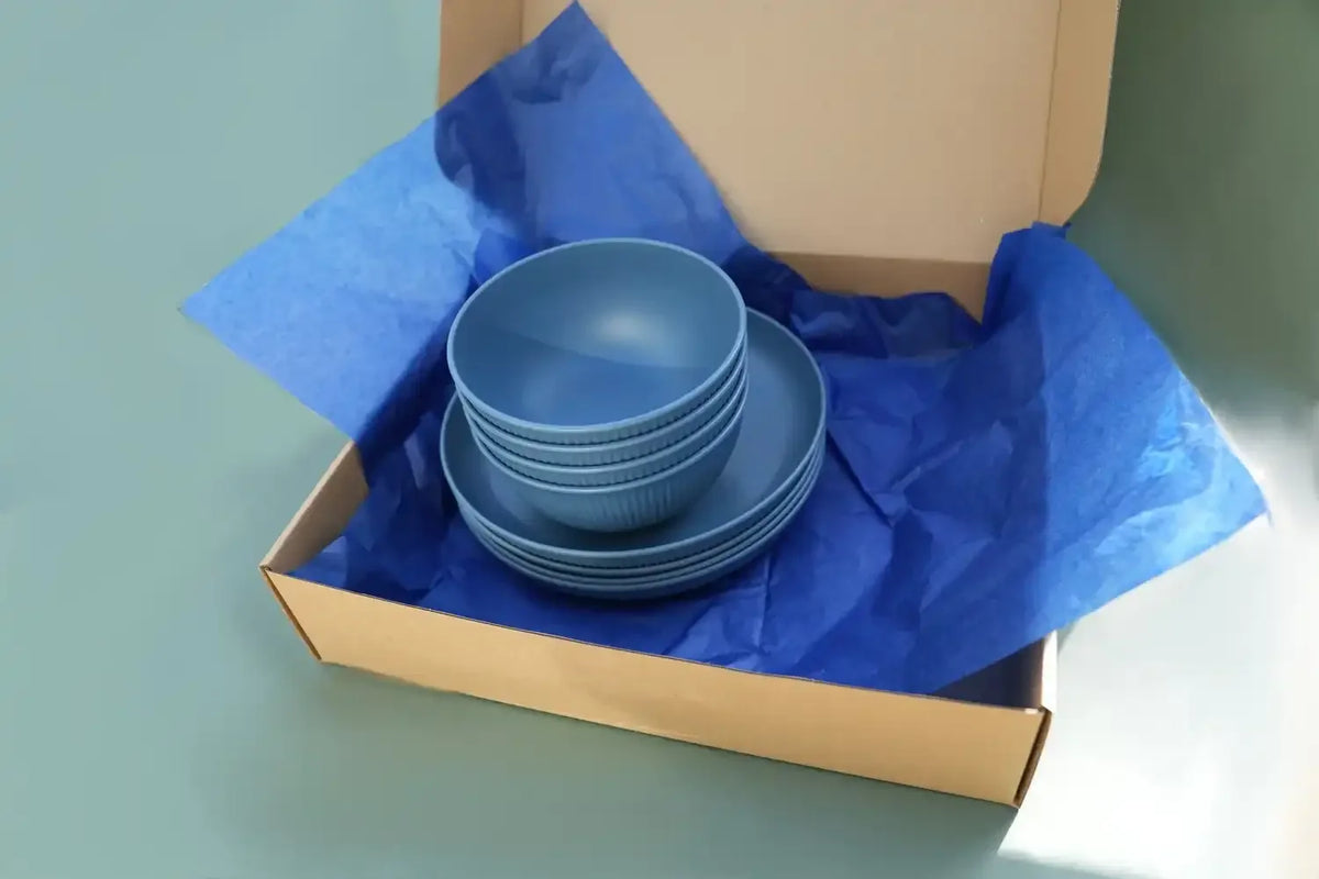 A stack of blue ceramic bowls and plates presented inside an open brown cardboard box lined with bright blue tissue paper, set against a solid pale green background.
