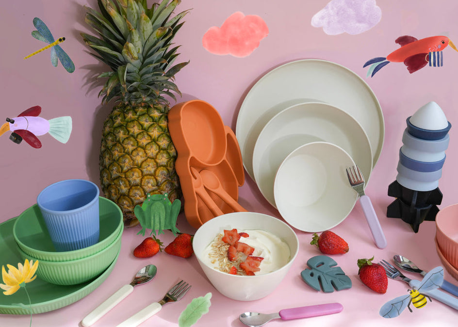 A styled flat-lay of colorful children's tableware on a pink background with whimsical paper cutouts of a pineapple, fish, dragonfly, clouds, and a rocket. The set includes plates, bowls, cups, spoons, and cutlery in pastel and bright colors, arranged around props like a strawberry bowl and a toy rocket.