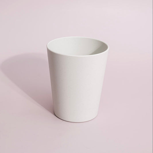 A minimalist white ceramic cup with a tapered cylindrical shape, standing on a light pink background with a soft shadow to the left.
