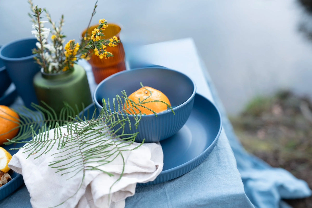 A styled tabletop scene with blue ceramic bowls and plates, a green vase with yellow flowers, orange fruit, and a white cloth, photographed in a real outdoor setting with a blurred natural background.