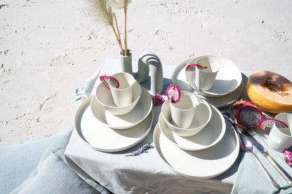 A styled outdoor dining setup featuring a set of white ceramic plates, bowls, and cups with pink dragon fruit garnish, arranged on a light blue linen cloth with a white textured wall background.