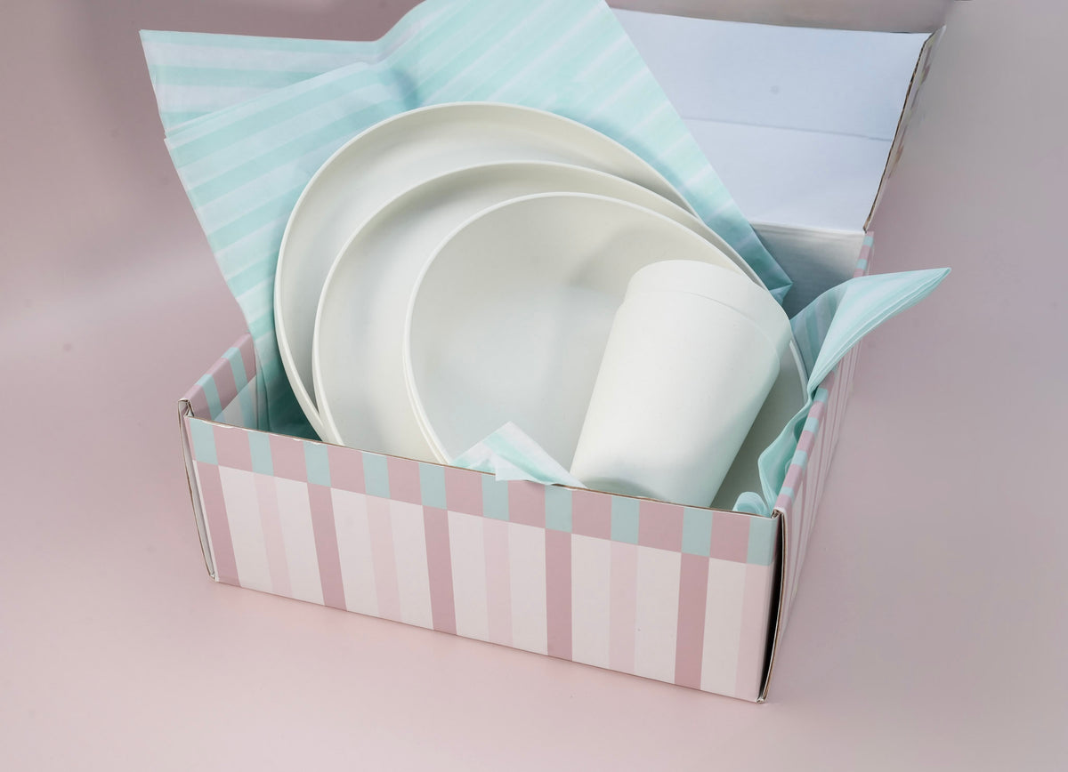A boxed set of white ceramic tableware, including plates and a cup, nestled in pastel striped tissue paper inside a pink and white striped gift box on a soft pink background.