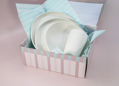 A boxed set of white ceramic tableware, including plates and a cup, nestled in pastel striped tissue paper inside a pink and white striped gift box on a soft pink background.