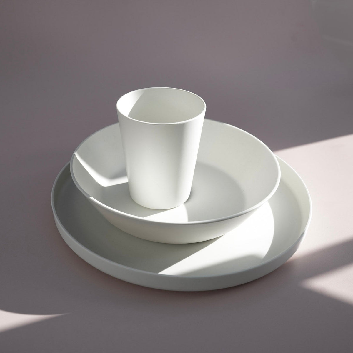 A minimalist white ceramic tableware set consisting of a cup, a shallow bowl, a deeper plate, and a large platter, arranged on a soft mauve background with dramatic diagonal shadows.
