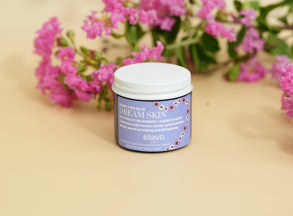 A small white jar of elavo Dream Skin cream sits on a beige surface with a soft-focus background of pink flowers.