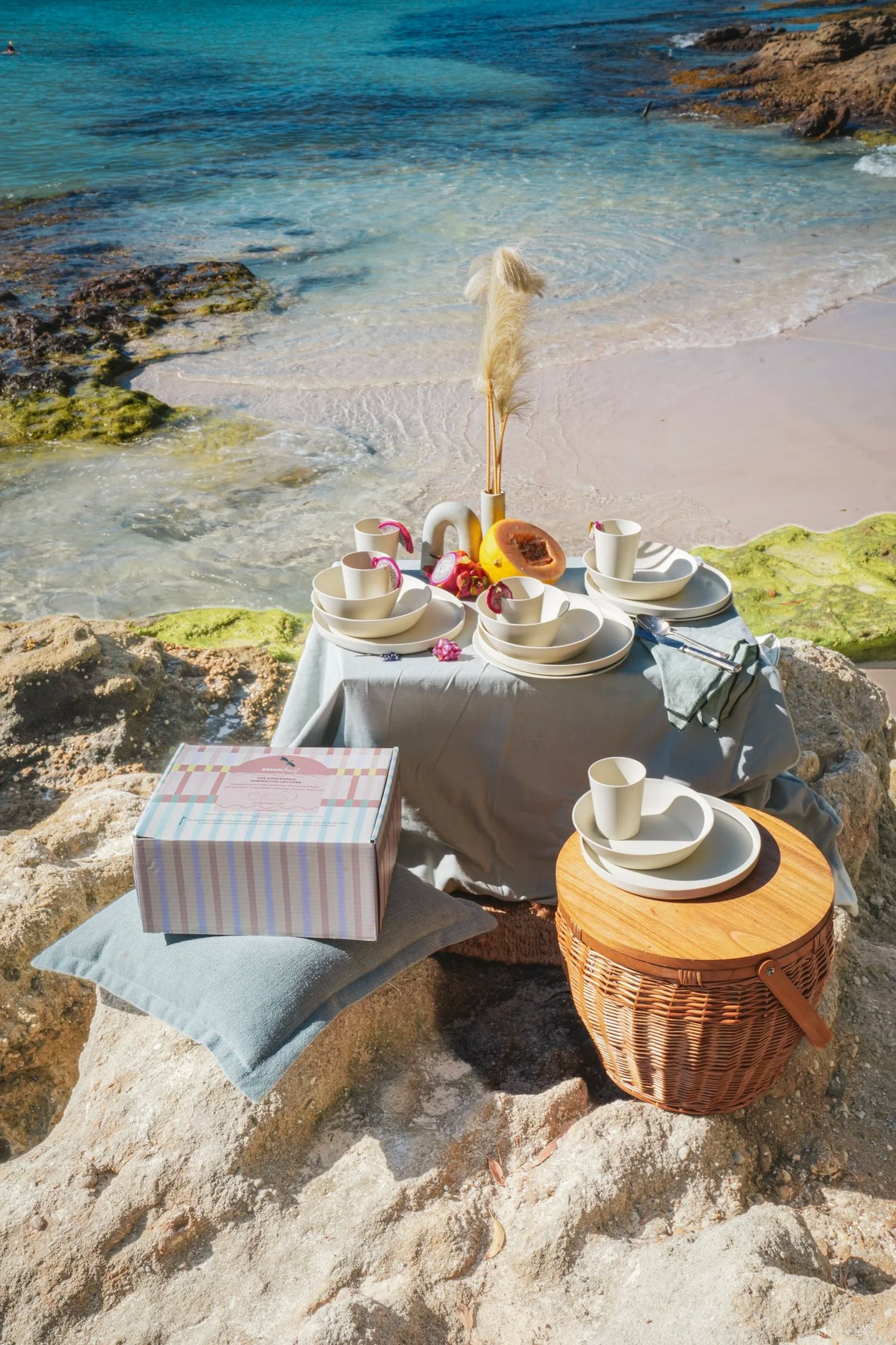 A styled beach picnic setup on rocky shorelines with a small table covered in a light gray cloth, white teacups and saucers, a striped gift box, a wicker picnic basket, and a pampas grass centerpiece, overlooking turquoise ocean water and pale sand.