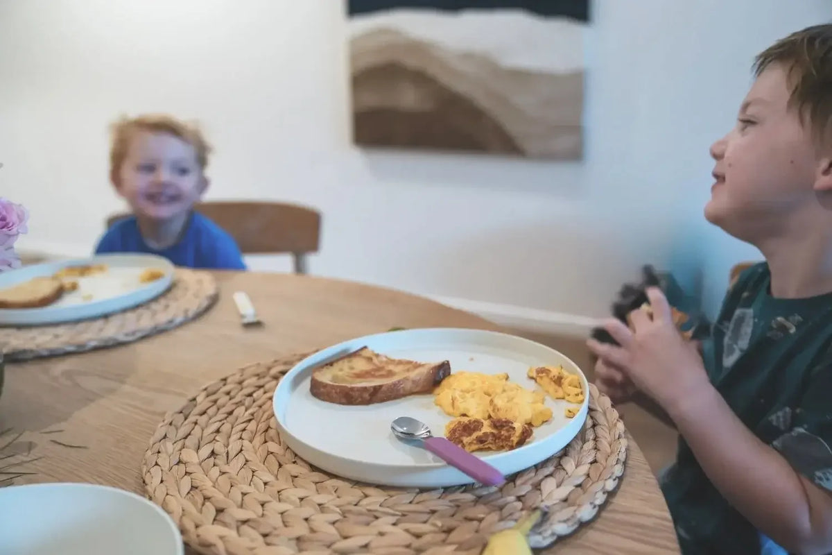 A lifestyle scene of a breakfast table with two children. In the foreground, a child on the right looks up at a plate containing scrambled eggs, toast, and a pink spoon.