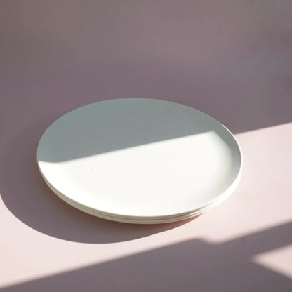 A stack of round white ceramic plates on a dusty pink surface with a diagonal shadow cast across the scene.