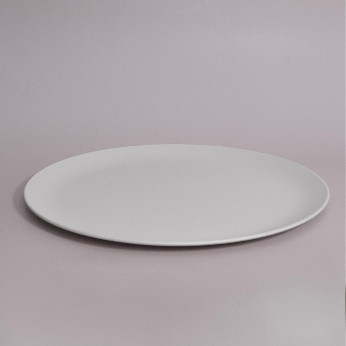 A minimalist white oval ceramic plate with a shallow rim, photographed against a seamless light gray studio background.