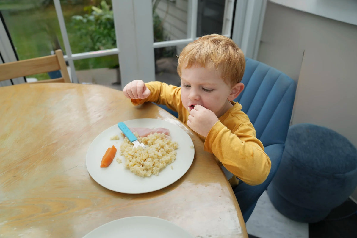 A young child with red hair sits in a blue chair at a wooden dining table, eating rice from a white plate with a blue-handled utensil. The child is wearing a yellow long-sleeve shirt in a home dining setting.