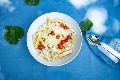 A top-down view of a white bowl filled with rotini pasta, topped with shaved cheese and cherry tomatoes, set on a blue tablecloth with green leaves and a blue napkin holding silverware.