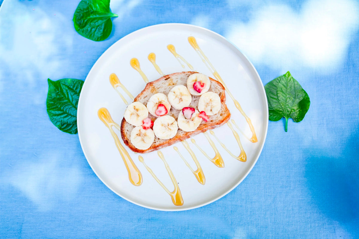 A top-down view of a white plate holding a slice of toast topped with banana slices, small red fruit pieces, and drizzles of honey, set on a blue fabric background with green leaves.