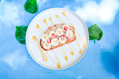 A top-down view of a white plate holding a slice of toast topped with banana slices, small red fruit pieces, and drizzles of honey, set on a blue fabric background with green leaves.