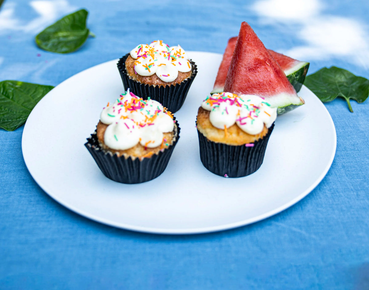Three small cupcakes with white frosting and colorful sprinkles on a white plate, served with watermelon slices, on a blue cloth background with green leaves.