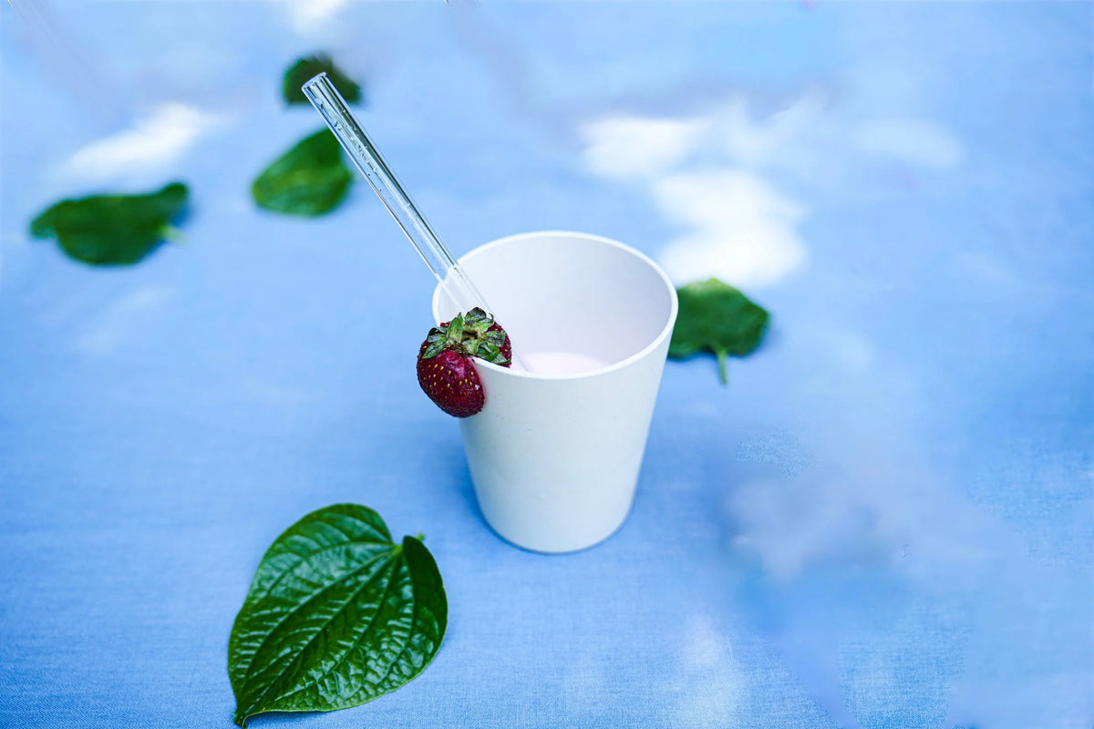 A white paper cup filled with a pale pink beverage, with a clear straw and a strawberry resting on the rim, set on a blue surface with scattered green leaves.