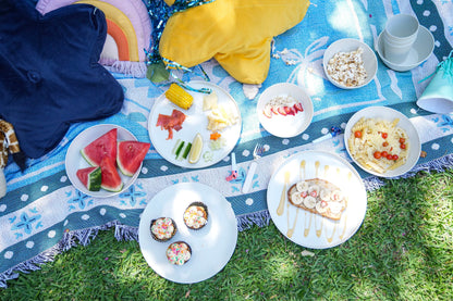 An overhead outdoor picnic scene on a blue patterned blanket spread over green grass, with plates of food including watermelon slices, popcorn, fruit, pasta, and small desserts. A navy top and a yellow blanket are also visible in the scene.