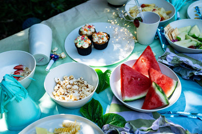 Outdoor picnic table setup with plates of watermelon slices, cupcakes, popcorn, sliced fruit, cups, and napkins on a light blue tablecloth.