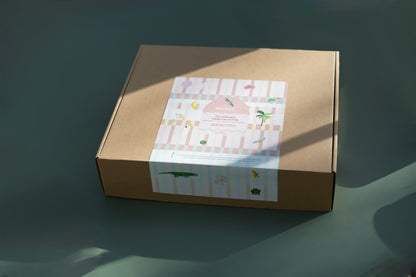 A square brown kraft paper box with a decorative pastel label on the front, photographed on a dark teal surface with soft diagonal shadows.