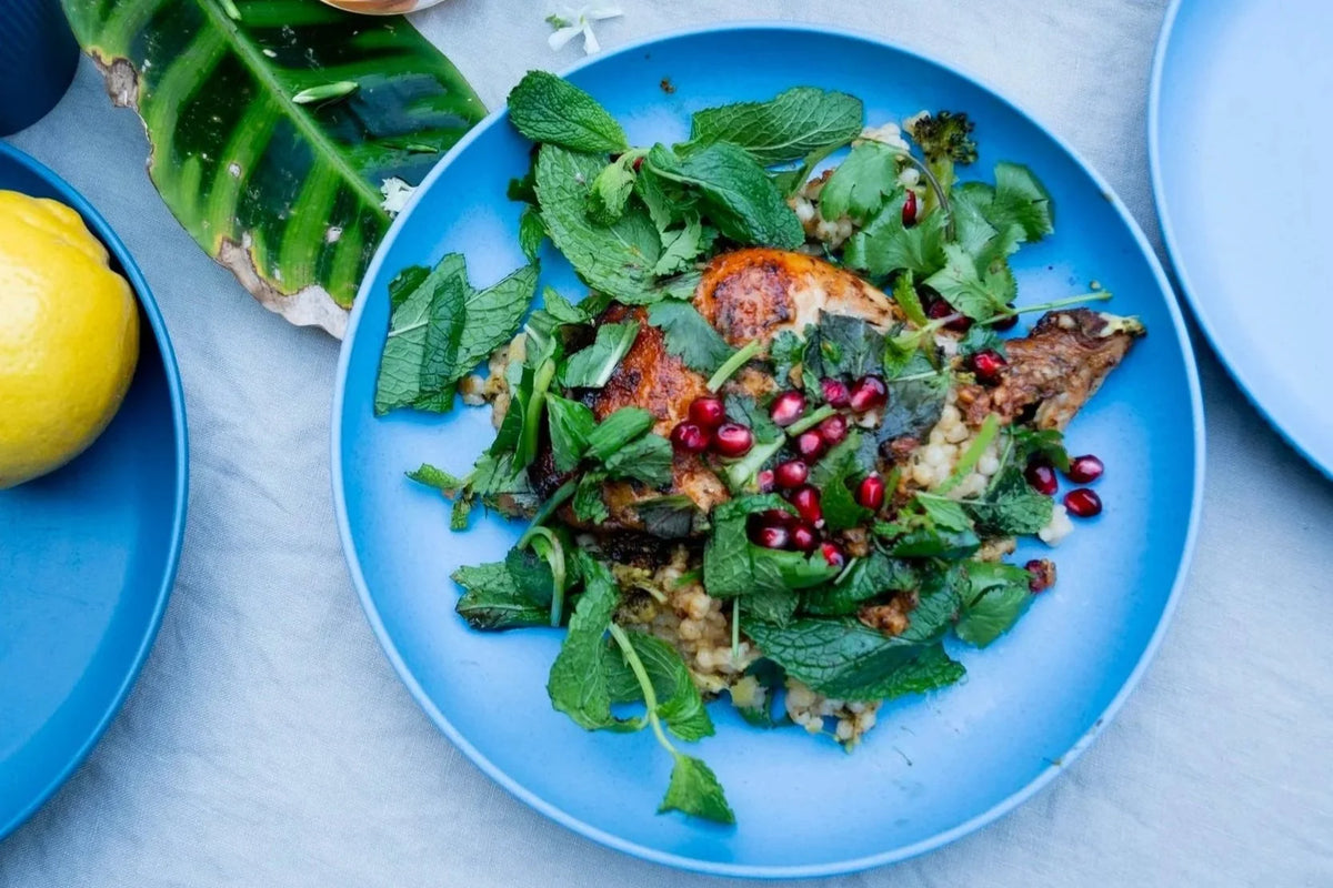A plated dish of cooked chicken with mint leaves, pomegranate seeds, and couscous on a blue plate, styled on a light gray surface with a lemon and tropical leaves nearby.