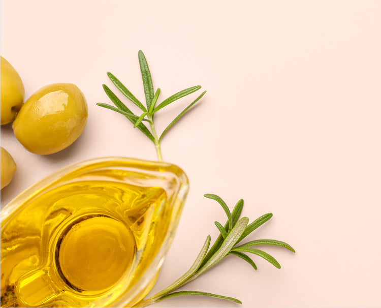 A glass bottle filled with golden olive oil, styled with whole green olives and rosemary sprigs on a soft pale pink background.