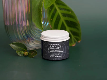 A small black skincare jar with a white lid labeled 'ELAVANT RESOLVE CLEANSING FACE BATH' sits on a dark green surface with green leaves and a ribbed iridescent glass vase in the background.