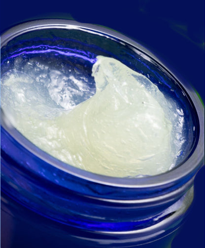 A close-up of a translucent pale yellow cosmetic balm or wax inside an open cobalt blue glass jar, set against a dark blue background.
