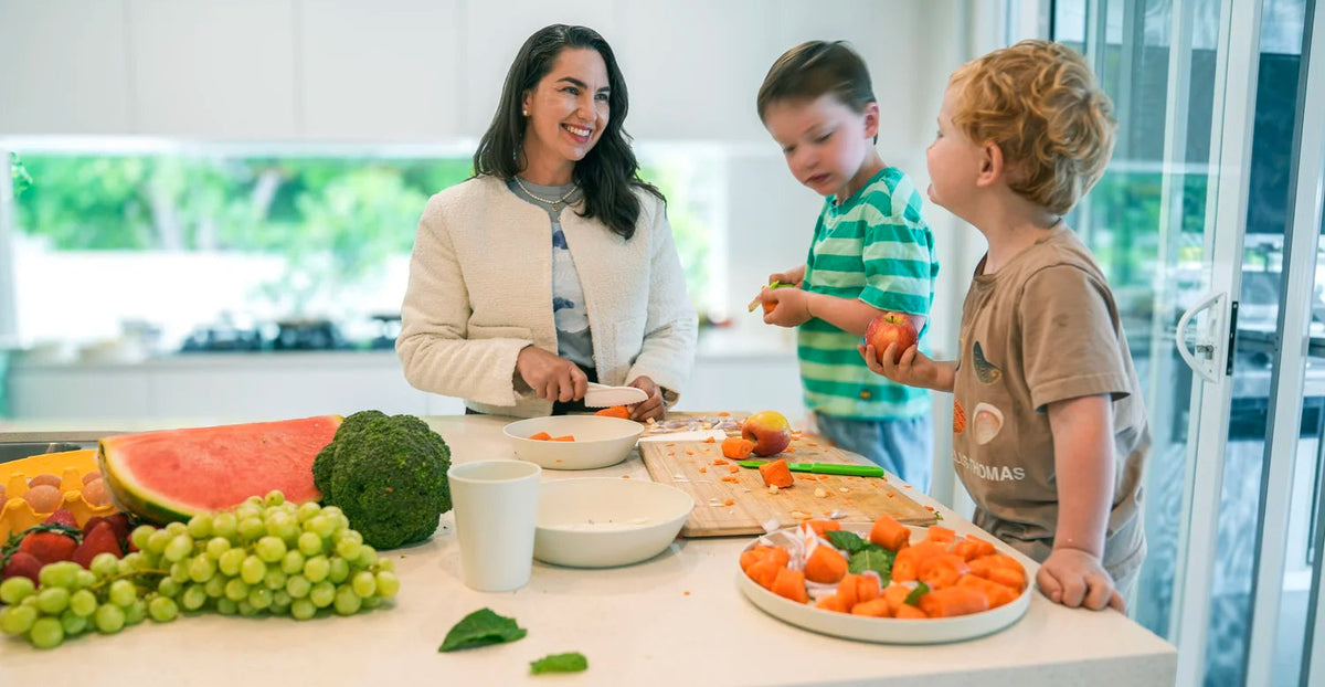 A woman and two young boys prepare food together in a bright modern kitchen. The kitchen island is covered with fresh fruits and vegetables, including watermelon, grapes, broccoli, and sliced carrots, with the boys holding pieces of fruit.