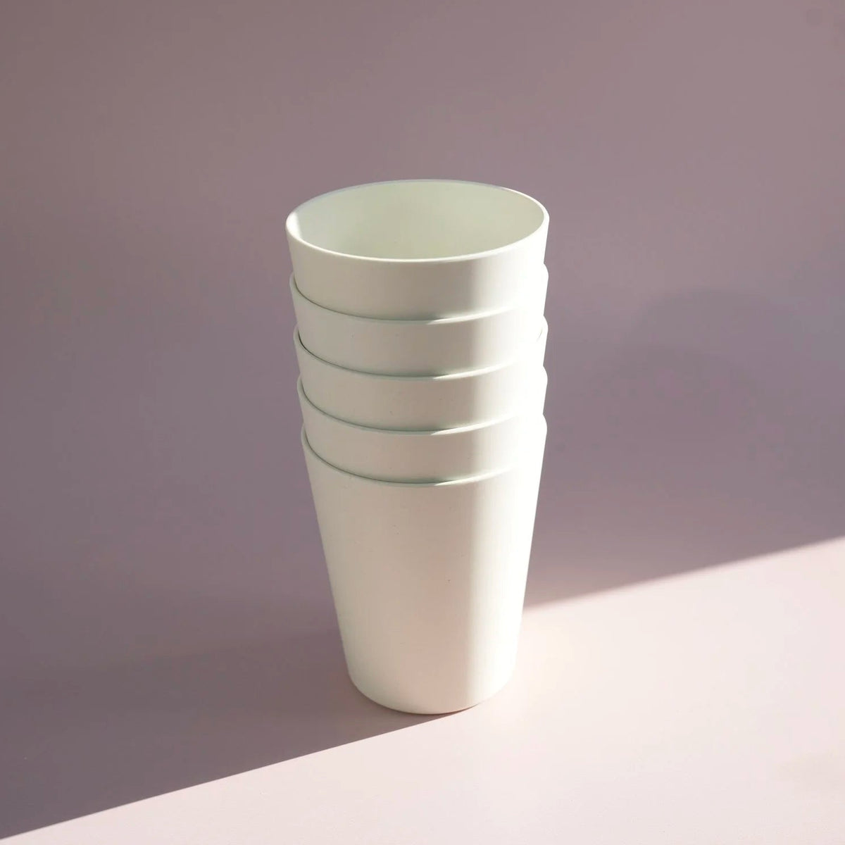 A stack of four matte white tapered cups with subtle mint green horizontal stripes near the rims, standing on a light surface against a soft lavender-gray background.