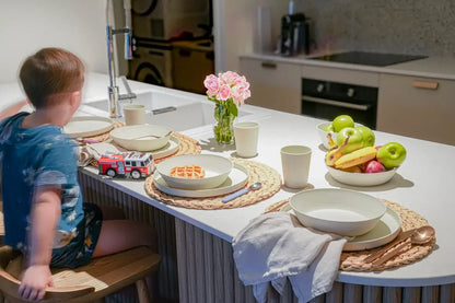 A young child wearing a blue tie-dye t-shirt sits on a wooden chair at a modern kitchen island, looking at a place setting with white ceramic plates, woven placemats, a small toy fire truck, and fresh fruit on a white countertop.