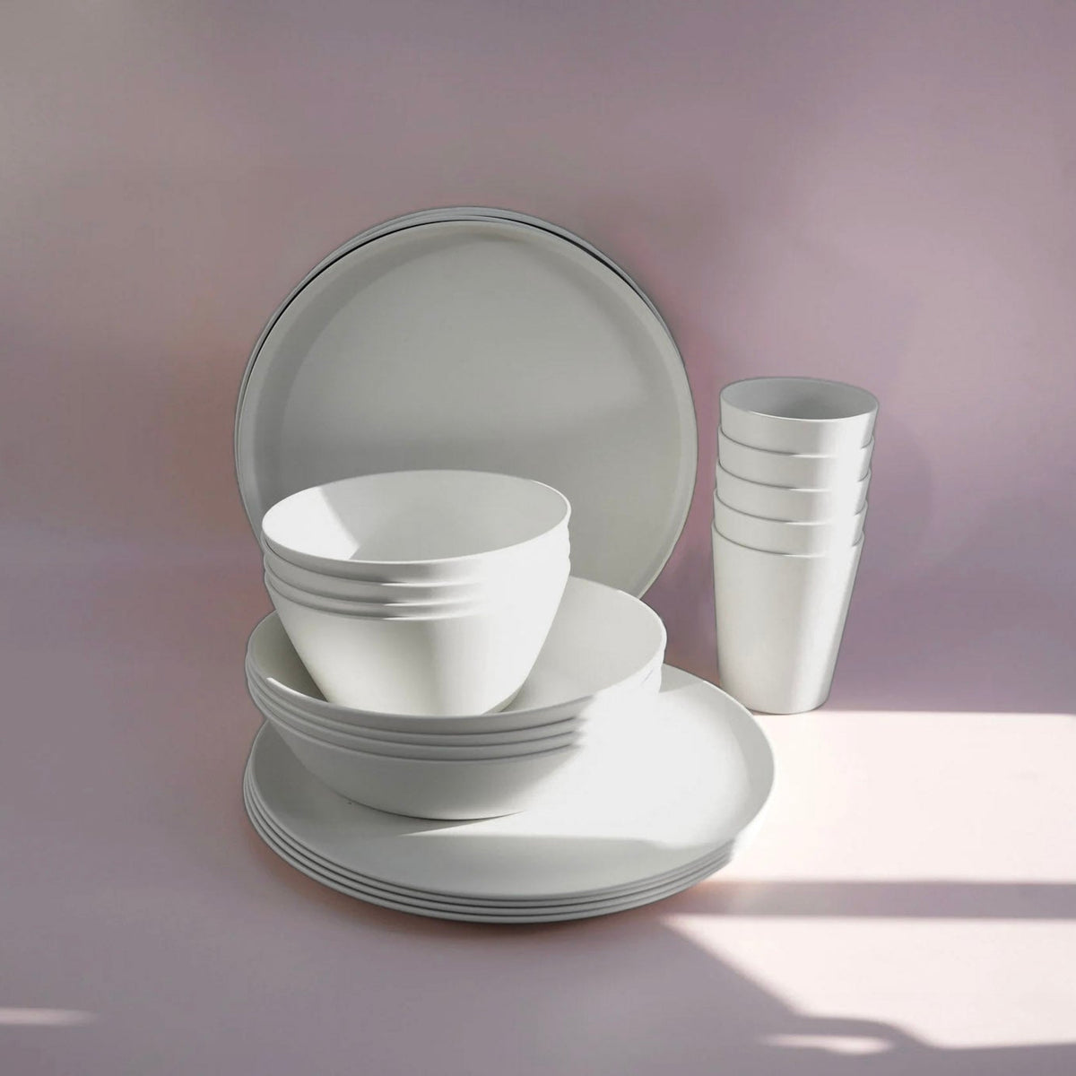 A minimalist white ceramic dinnerware set including plates, bowls, and cups, arranged on a soft pink surface with soft shadows.