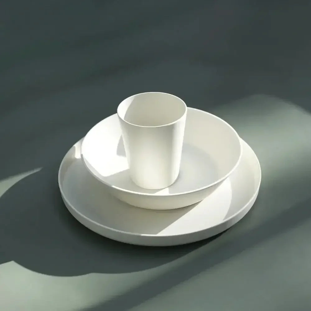 A minimalist white ceramic dinnerware set consisting of a shallow plate, a bowl, and a matching cup, arranged in a stack on a dark gray surface with dramatic side lighting.