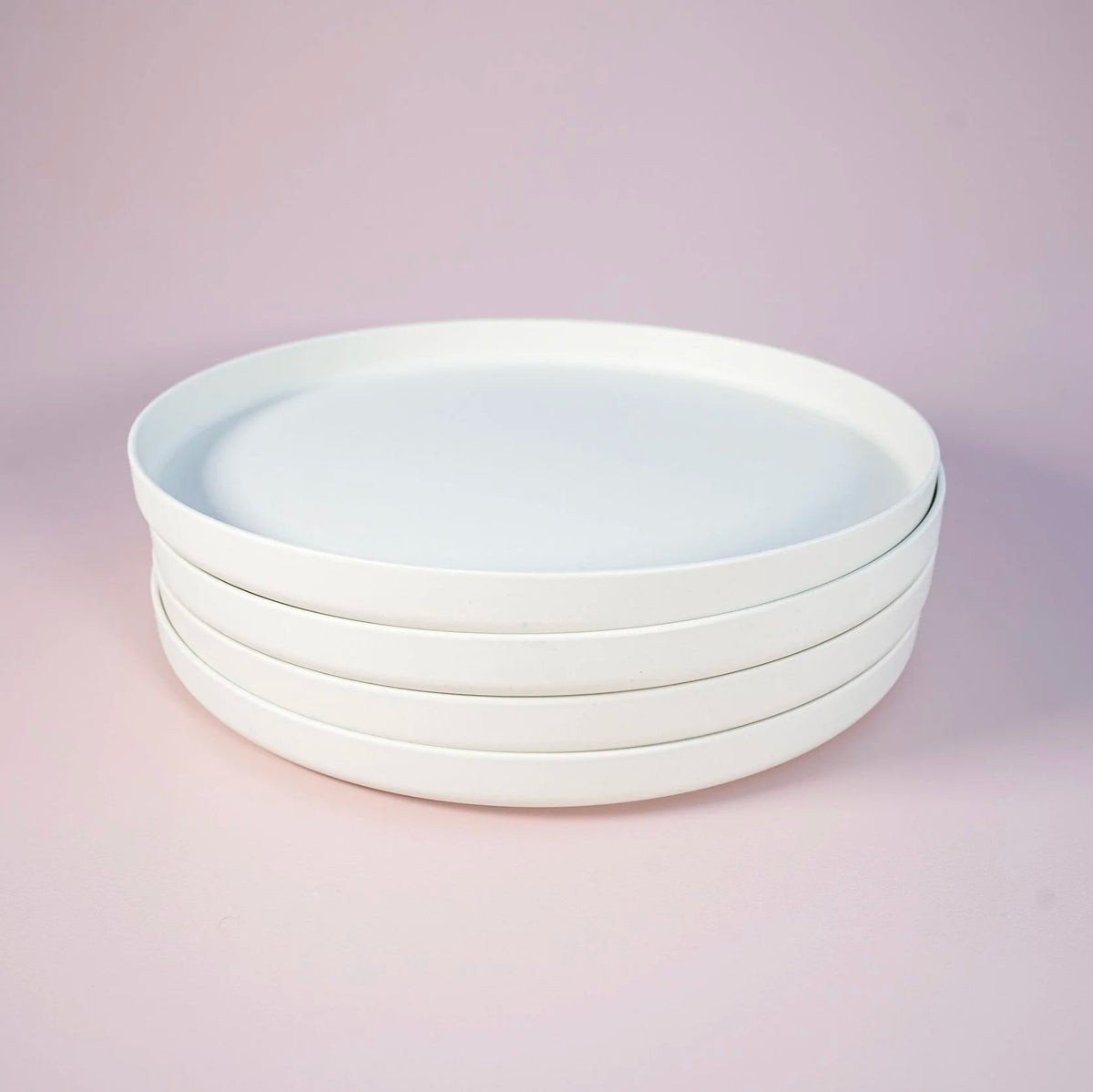 A stack of four round white ceramic looking platic plates on a soft pink backgrond