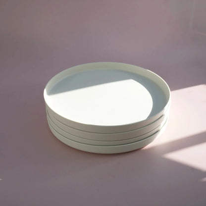 A stack of four minimalist white ceramic look plastic plates with a shallow, wide rim, photographed on a matching white surface under soft, directional sunlight that casts shadows.