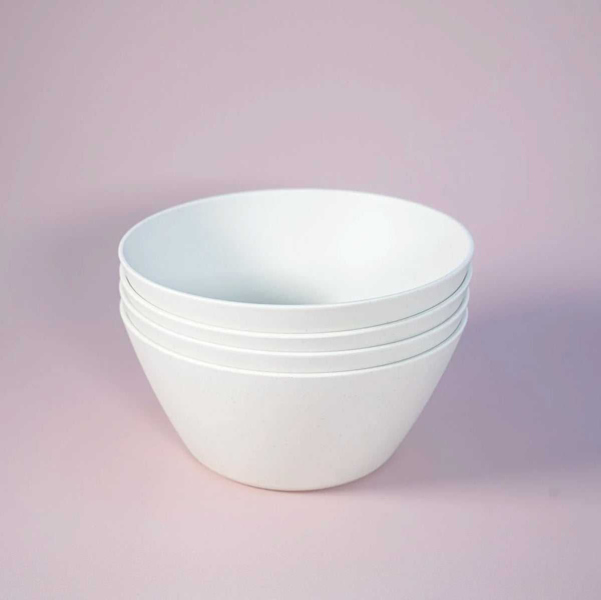 A stack of four matte white ceramic bowls with a wide, rounded shape, set against a solid light pink background.