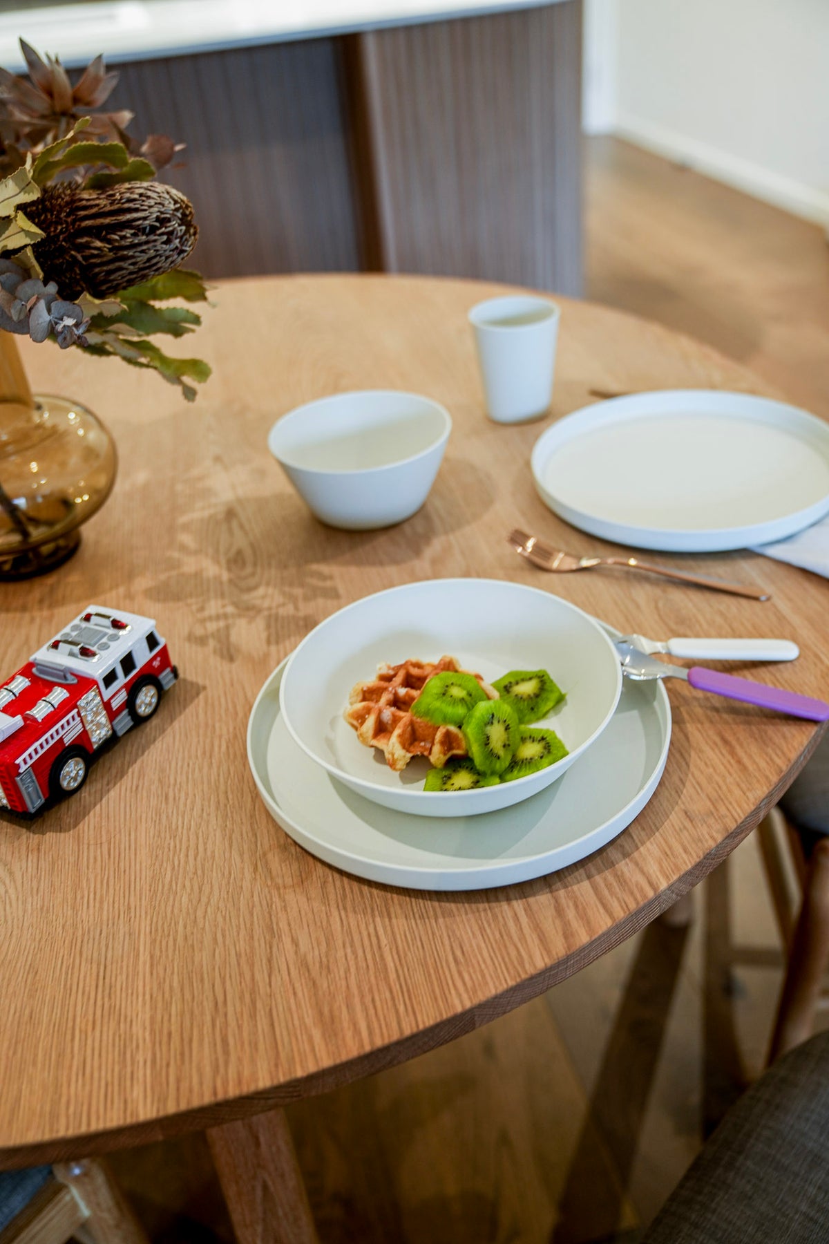 A styled dining table scene with white ceramic bowls, plates, cutlery, a small cup, and a red toy fire truck on a light wood table. No person or animal is present, but this is not a clean isolated product shot; it is a real interior environment, so subject_type is Lifestyle.