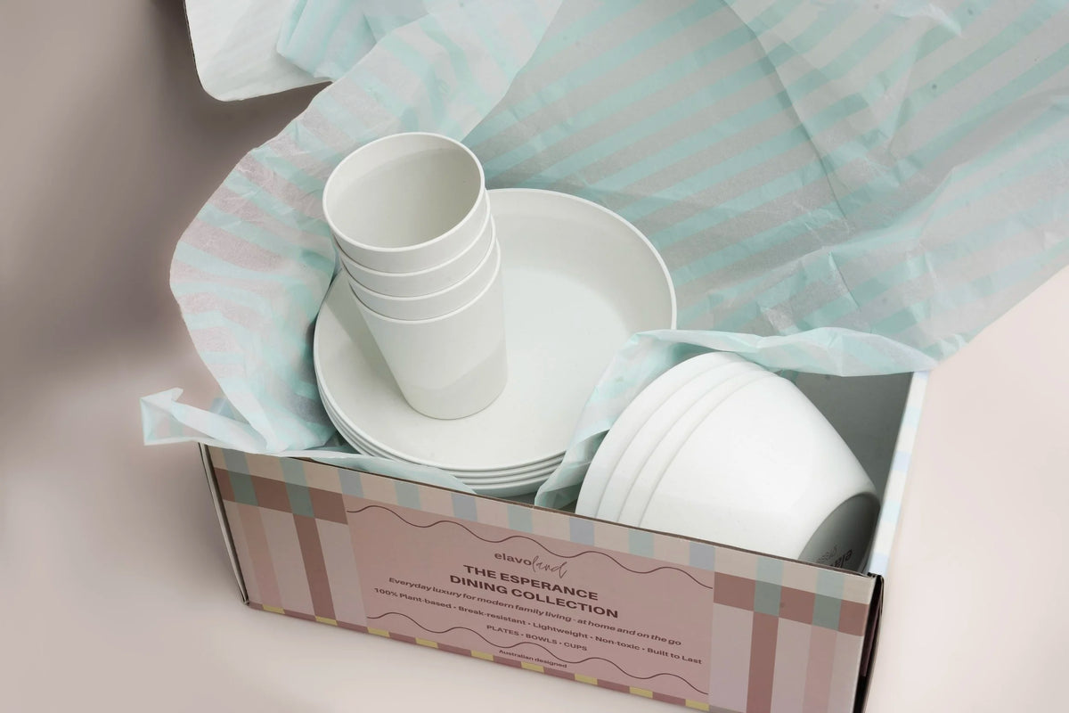 A boxed dinnerware set from The Esperance Dinner Collection, featuring white ceramic cups, plates, and bowls nestled in pastel striped tissue paper inside a patterned gift box.