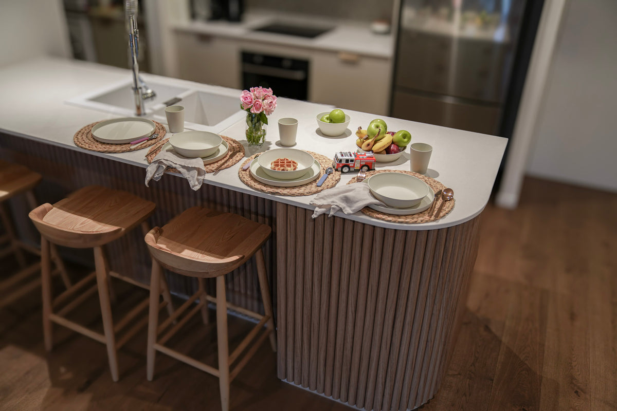 A modern kitchen island with a fluted wood-look base, white countertop, and three wooden bar stools. The island is styled with plates, placemats, a small vase of pink flowers, and fruit, set in a bright kitchen with wood floors.
