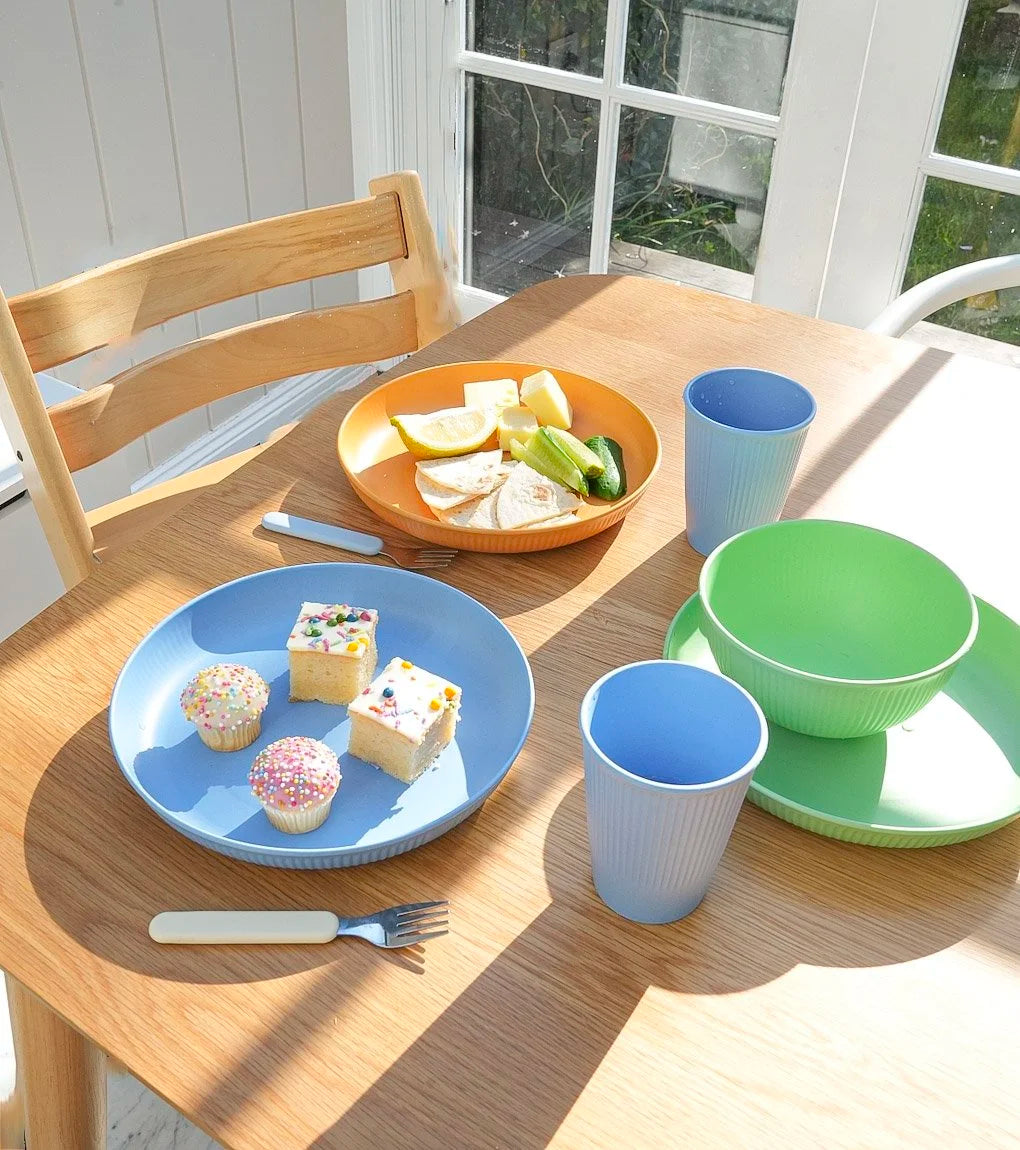 A set of pastel-colored children's tableware including a blue plate with cupcakes, an orange plate with fruit, a green bowl and saucer, and matching blue and green cups, arranged on a wooden dining table with a chair and window in the background.