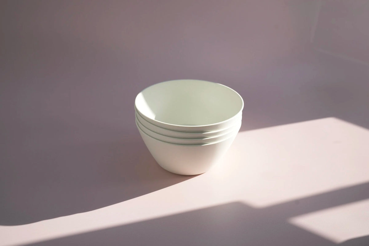 A stack of five white ceramic bowls with thin green stripes around the rims, set on a light pink surface with soft diagonal shadows.