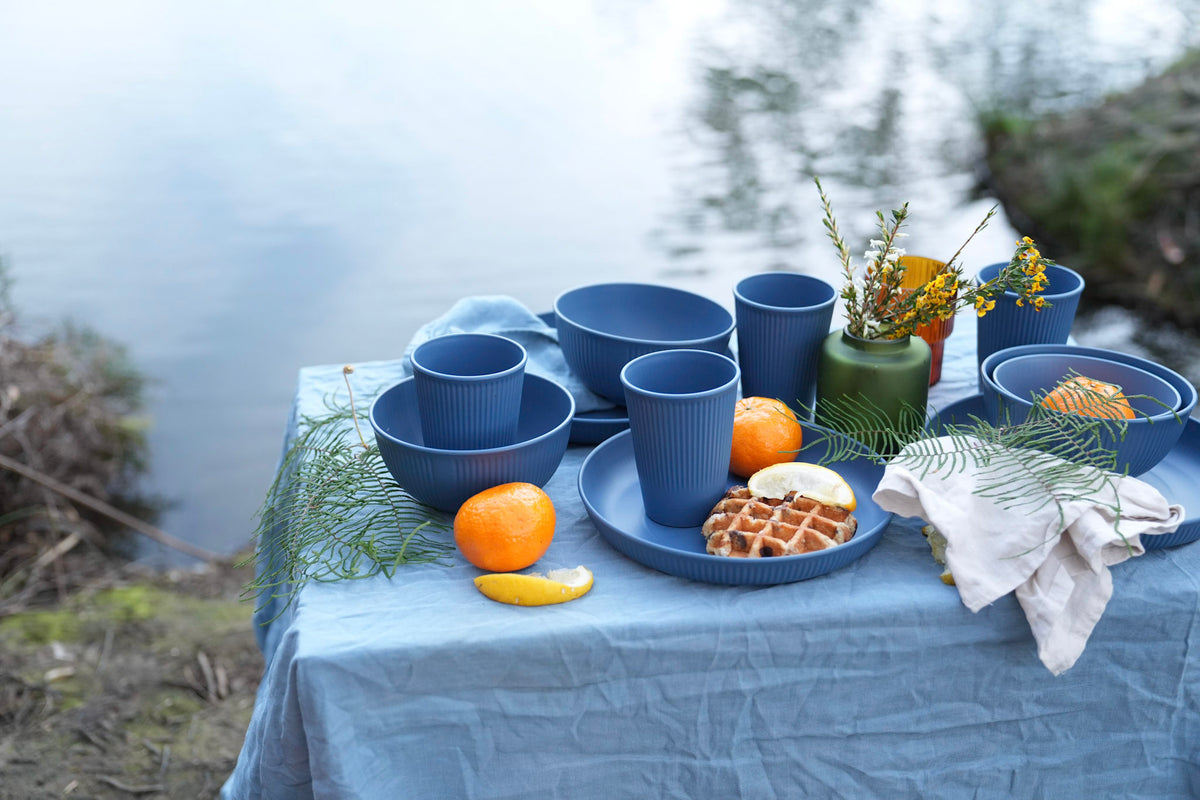 A styled outdoor dining setup by a lake or river, featuring a coordinated set of blue ceramic bowls, cups, and plates on a blue tablecloth, alongside oranges, waffles, and a small green vase with yellow flowers.