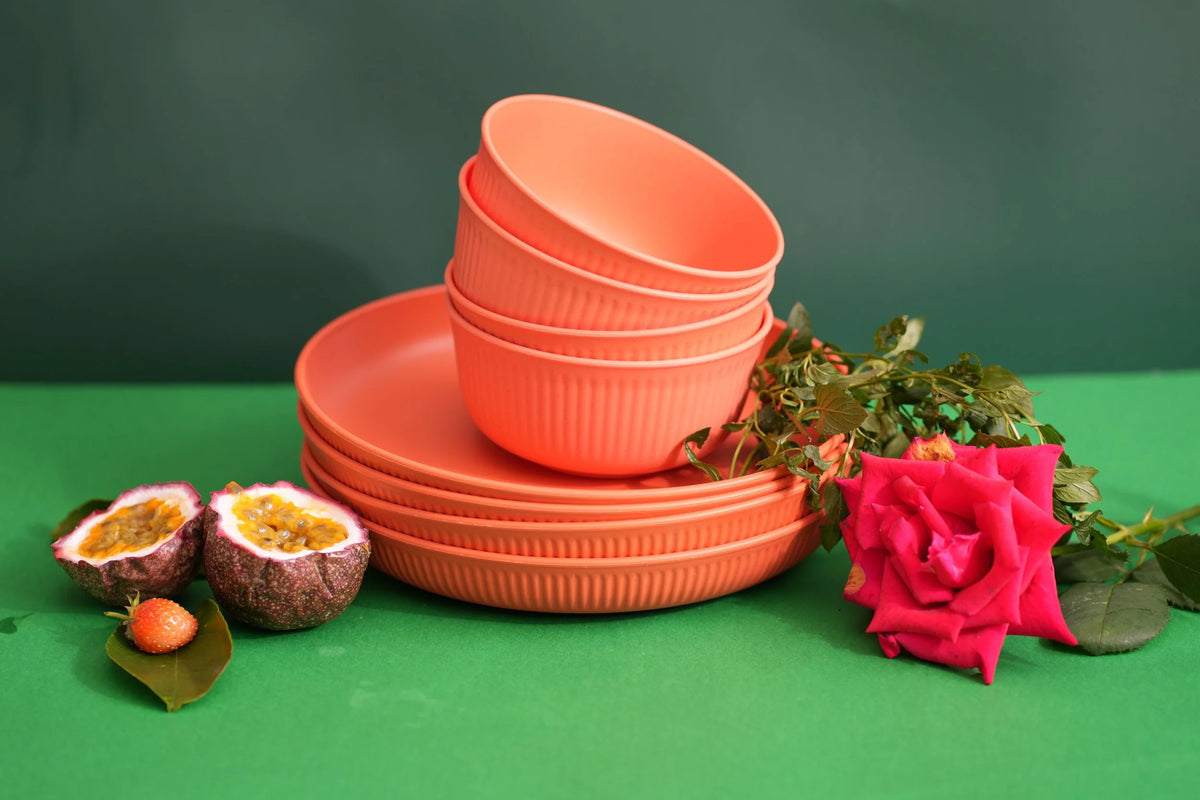 A set of coral orange plastic bowls and plates with a ribbed exterior, styled with a pink rose, halved passion fruit, and green leaves on a green surface against a dark green background.