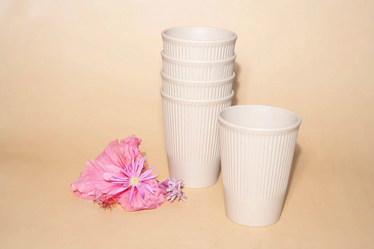 Three white ribbed disposable cups, including a stack of four and one single cup, displayed on a beige background with pink flowers in the foreground.
