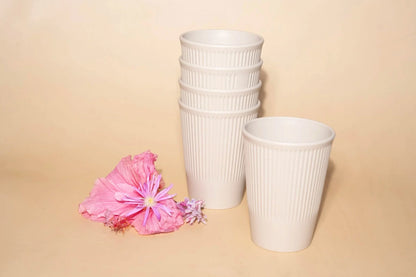 Three white ribbed disposable cups, including a stack of four and one single cup, displayed on a beige background with pink flowers in the foreground.