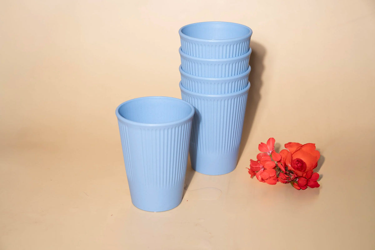 A set of light blue ribbed plastic cups, including one cup standing alone and a stack of five cups, displayed on a beige background with a small red flower prop.