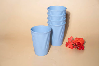 A set of light blue ribbed plastic cups, including one cup standing alone and a stack of five cups, displayed on a beige background with a small red flower prop.