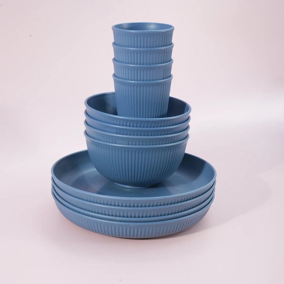 A set of blue ribbed dinnerware, including plates and bowls, stacked neatly on a light pink studio background.