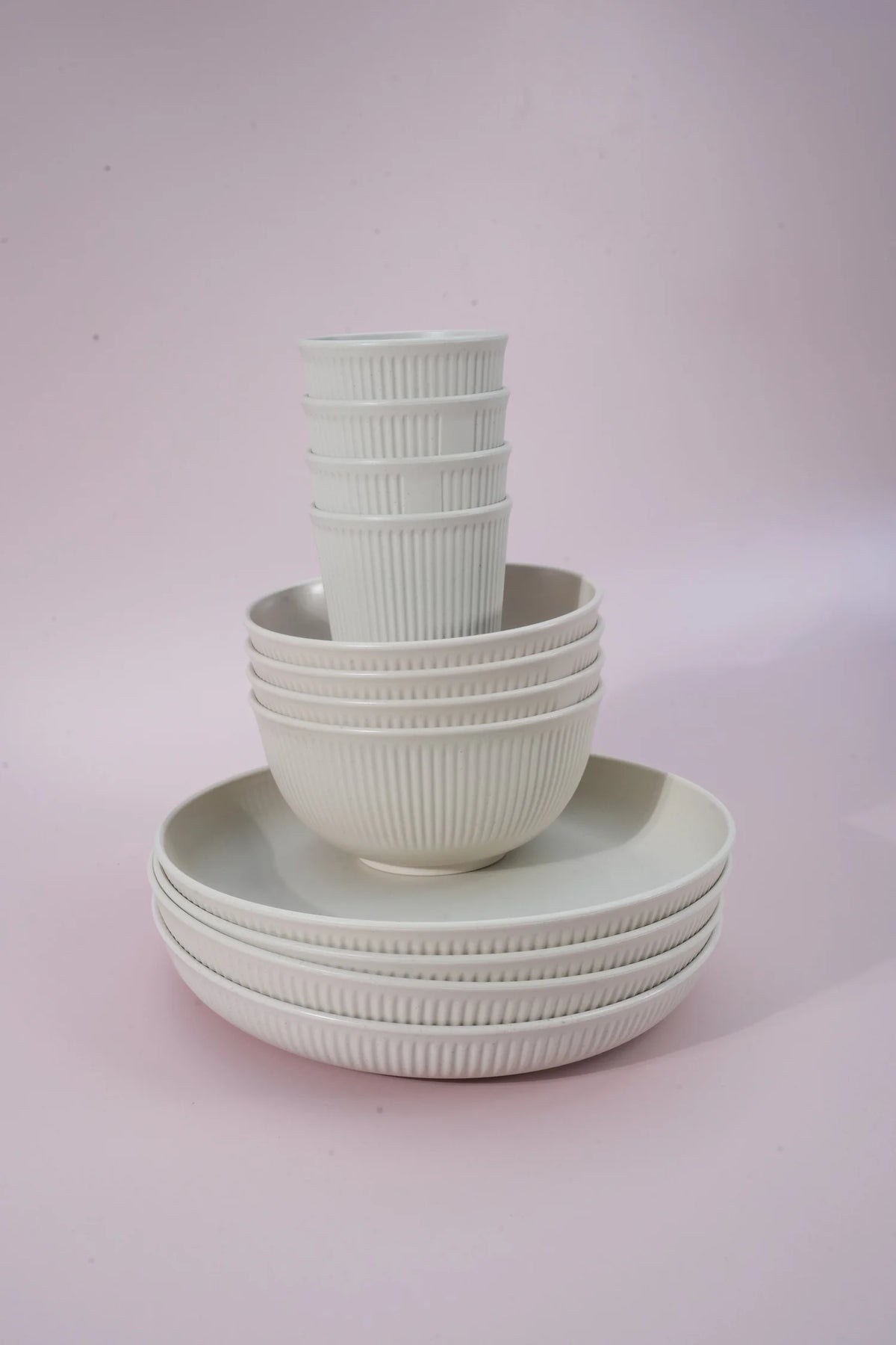 A set of ribbed ceramic tableware, including bowls and plates, stacked neatly on a soft pink studio background.